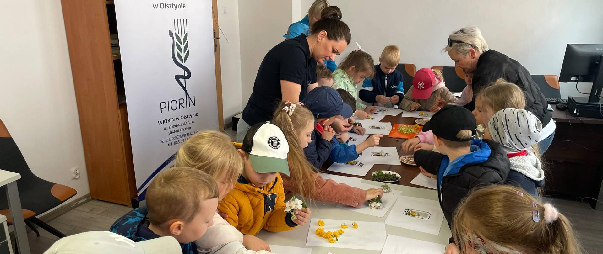 Grupa dzieci zebranych przy stołach przygotowuje słoiczki wspomnień, przyklejając żywe rośliny do kartonowych kartek z nadrukiem słoiczka. W tle widoczne jest logo PIORiN
