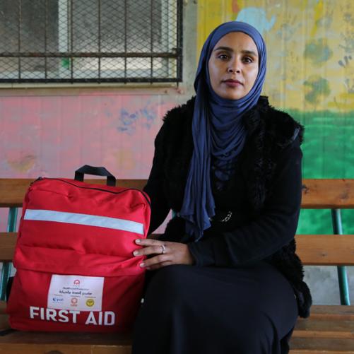 Kobieta w chuście z czerwonym plecakiem z napisem First Aid i logo PAH, Polska pomoc, Care