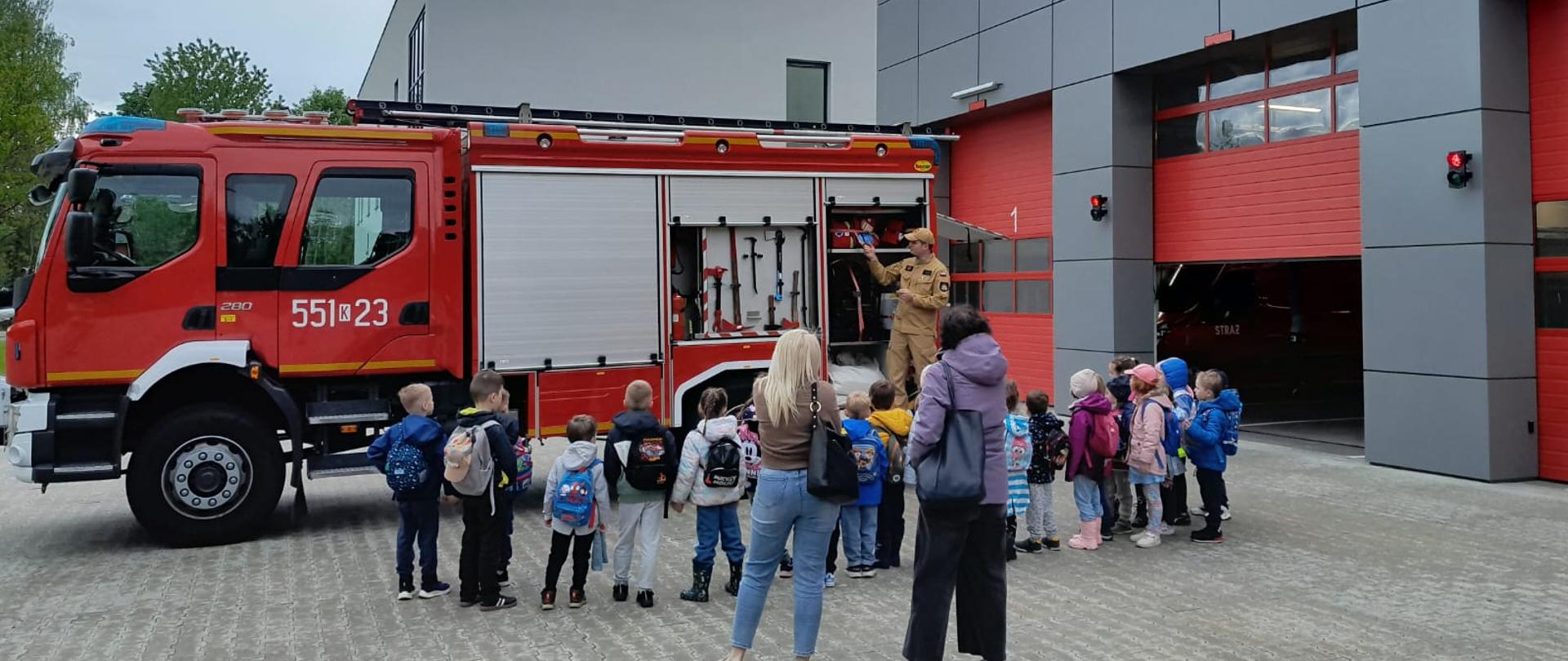 Na pierwszym planie widać dzieci oglądające sprzęt znajdujący się na wyposażeniu samochodu strażackiego. Na drugim planie znajduje się samochód strażacki z otwartymi skrytkami oraz strażak prezentujący znajdujący się na wyposażeniu samochodu sprzęt. 