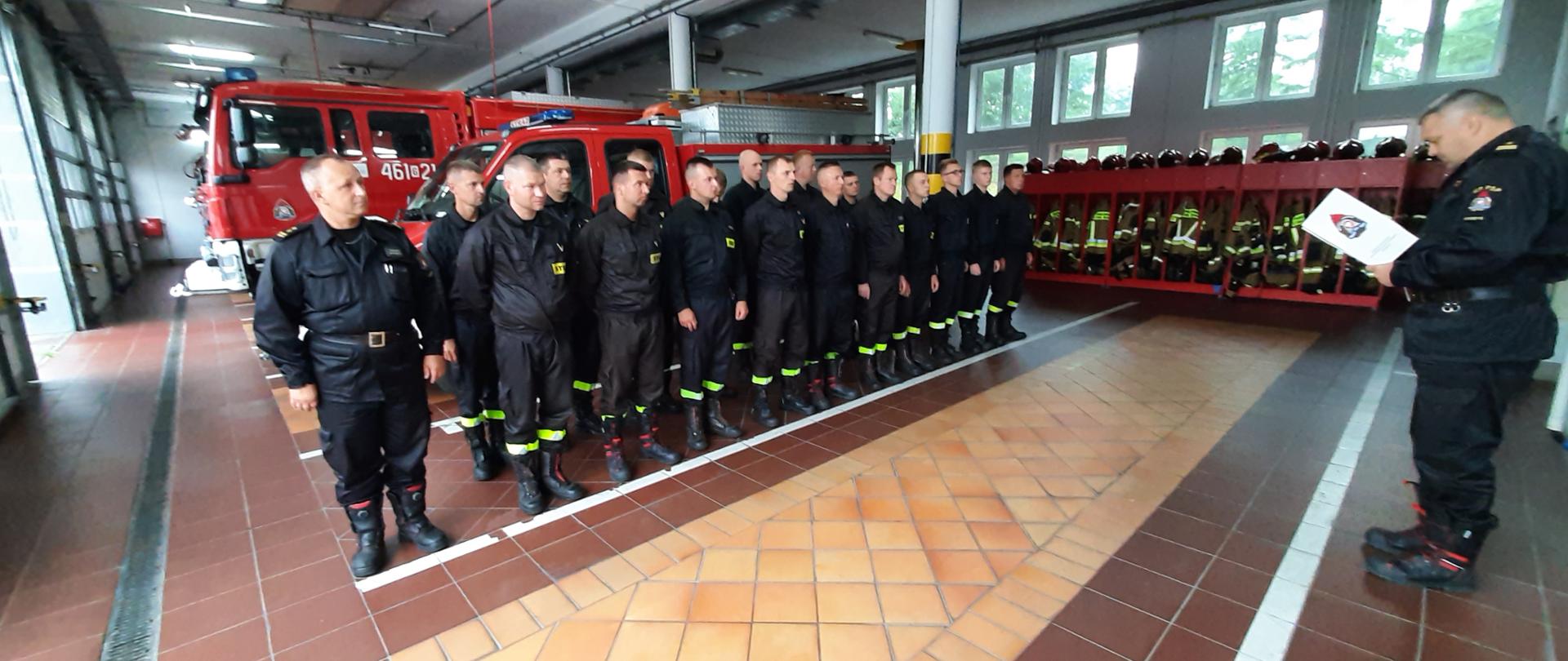 Komendant Powiatowy stoi przed zastępem strażaków podczas uroczystej zbiórki w garażu bojowym.