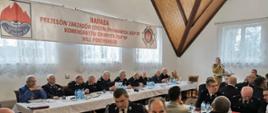 Strażak Państwowej Straży Pożarnej w mundurze piaskowym stoi za mównicą przemawiając do strażaków ochotniczych straży pożarnych siedzących za stołami na których rozłożone są butelki z wodą. 