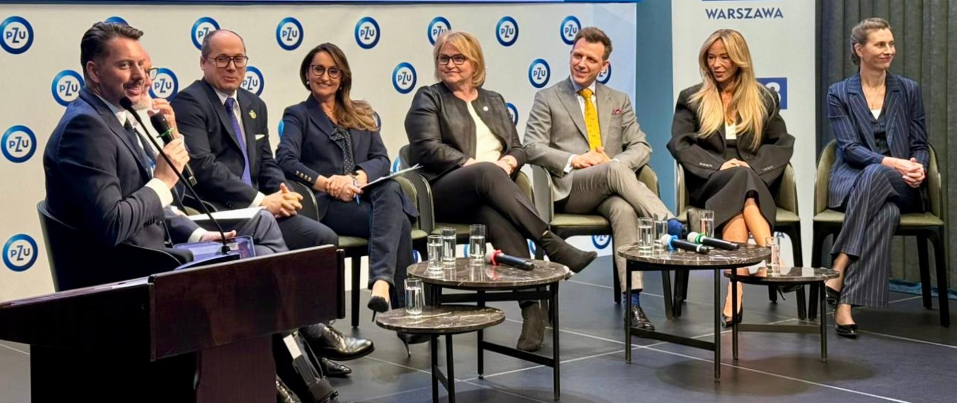 Paneliści na scenie, przed nimi stoi mównica i stoliki kawowe.