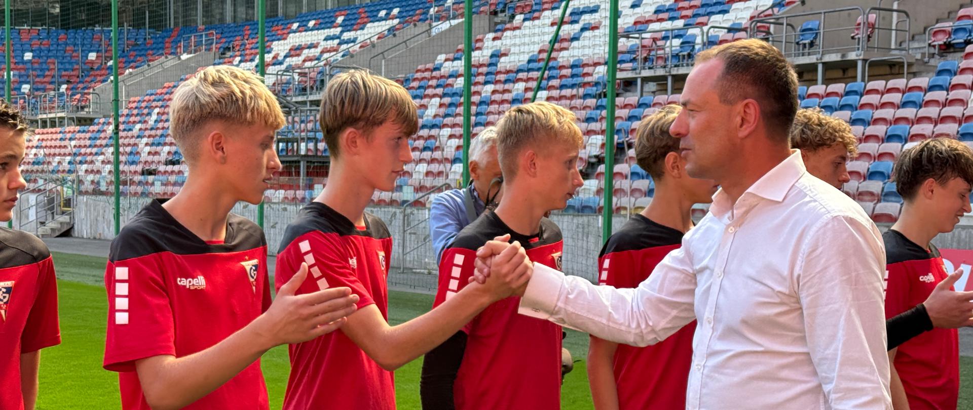Zdjęcie zrobione na stadionie piłkarskim. Chłopcy w czerwonych koszulkach i czarnych krótkich spodenkach stoją w szeregu. Minister Piotr Borys przechodzi przed nimi, podając każdemu rękę. W tle trybuny. 