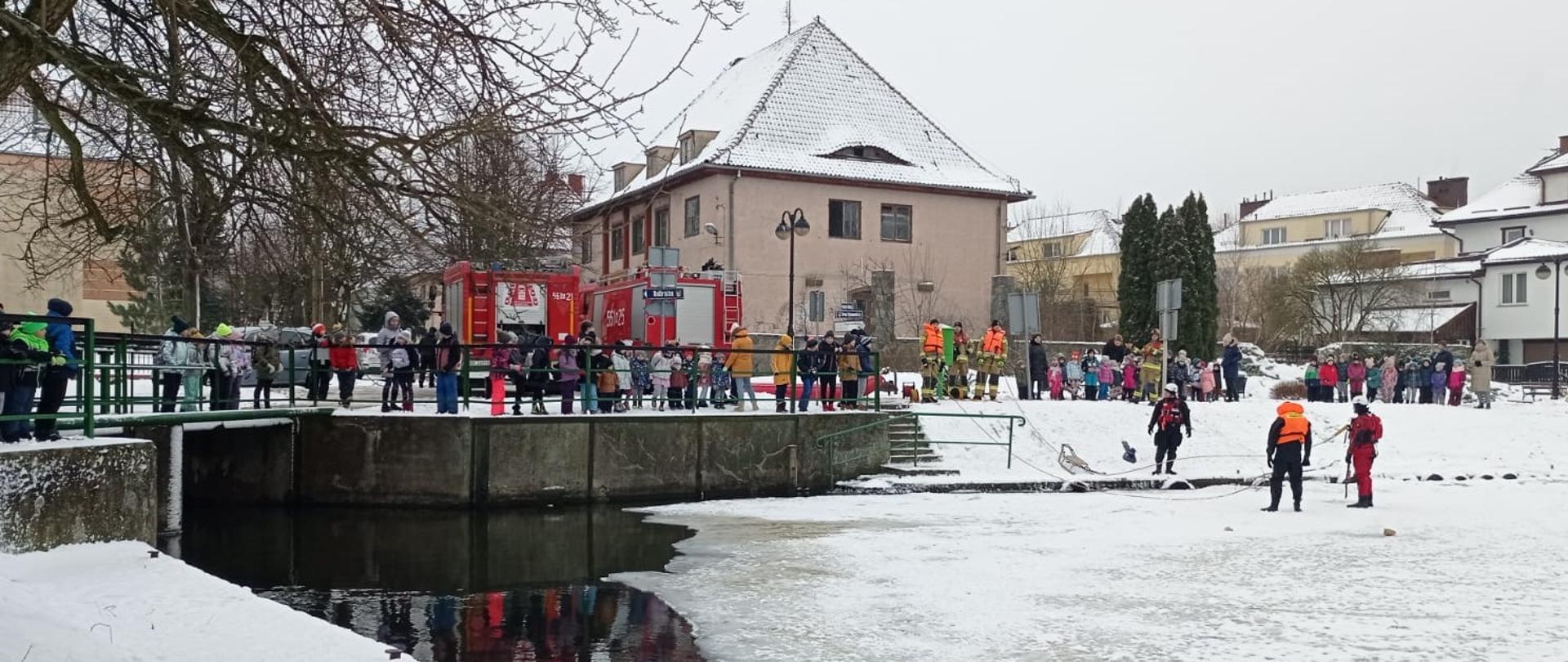 Na brzegu nad kanałem stoją dzieci, które oglądają pokazy z ratownictwa lodowego organizowane przez strażaków. 2 strażaków stoi na lodzie i omawia techniki ratowania osoby pod która załamał się lód. Za dziećmi w stoją dwa samochody gaśnicze i widoczne są bloki mieszkalne. 