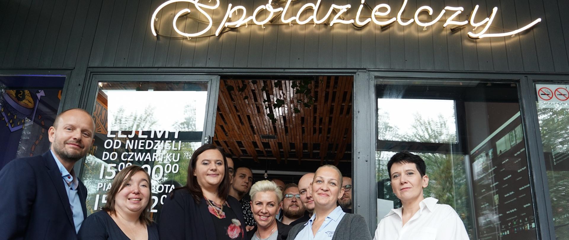 Grupa osób stoi przed lokalem spółdzielczy wśród nich minister Jarosińska-Jedynak 