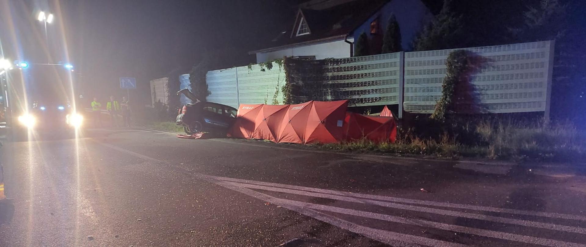 Zdjęcie przedstawia wypadek dwóch samochodów osobowych. Jedno z aut stoi częściowo na jezdni, częściowo w przydrożnym rowie. Drugie auto stoi za parawanem strażackim. Pora nocna. Na zdjęciu widać również samochód strażacki. 