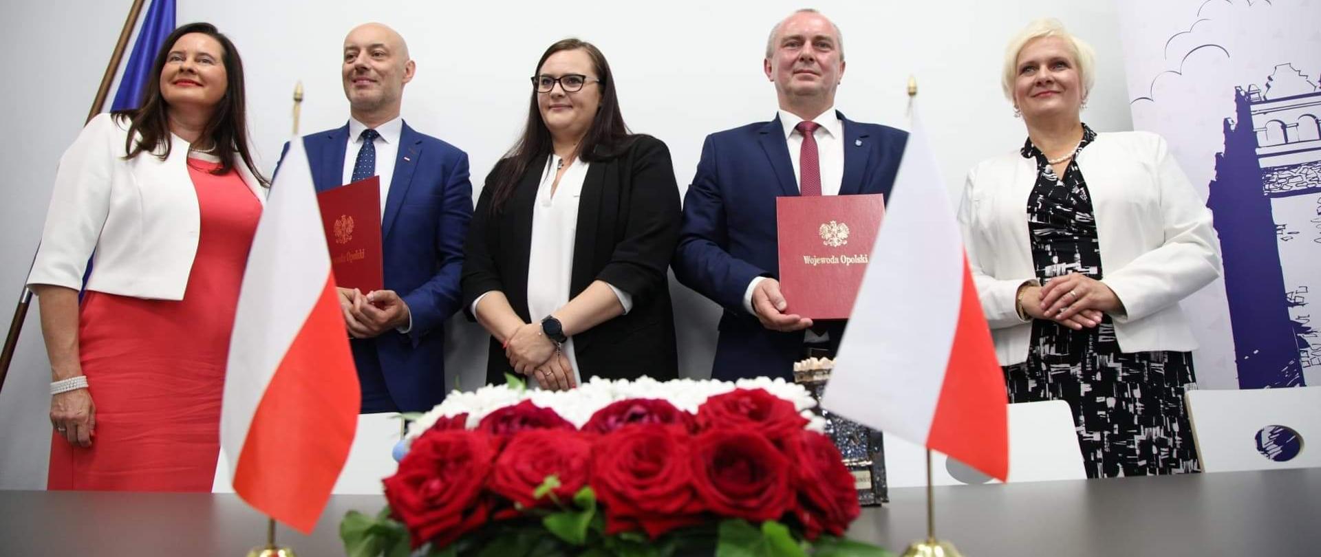 Na pierwszym planie stół, na którym po środku stoi wiązanka kwiatów a obok flagi Polski. Dalej stoi pięć osób. Pośrodku minister M. Jarosińska-Jedynak
