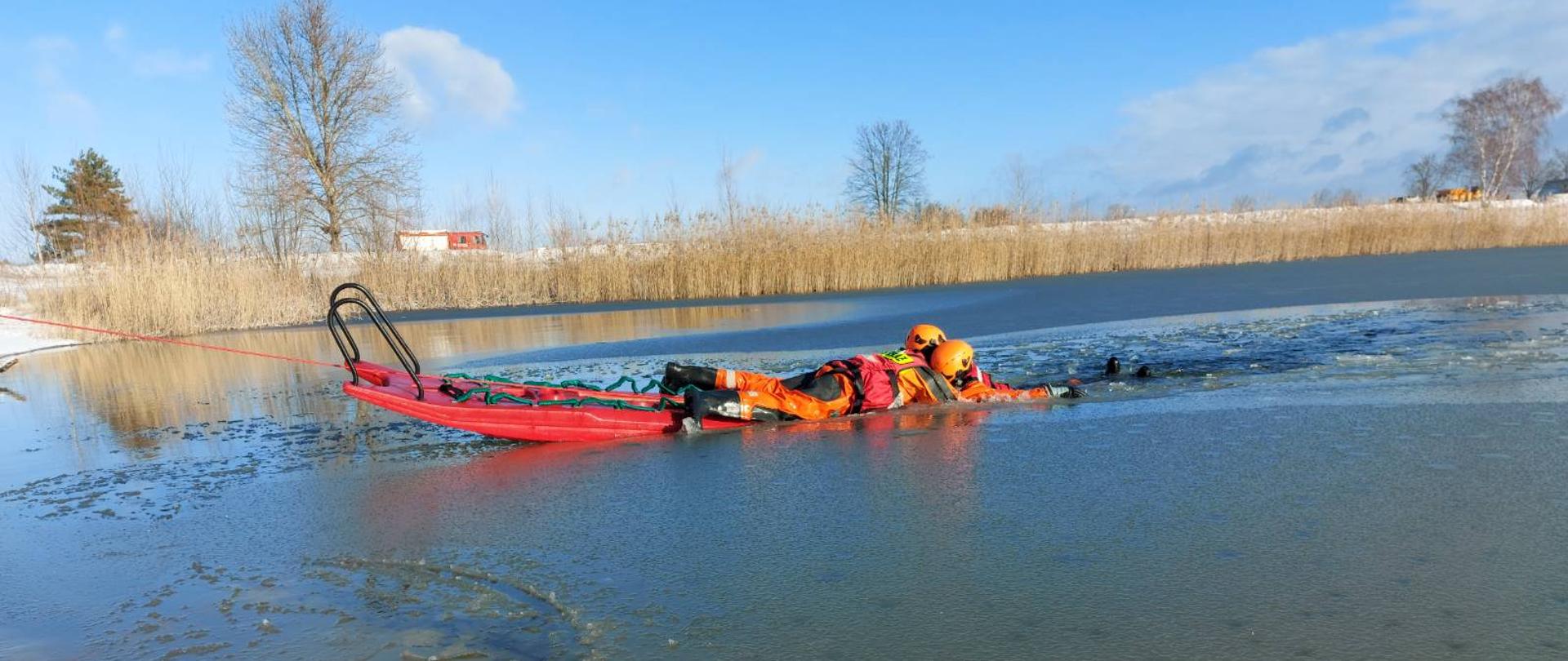 . Akwen w Sierakowie. Strażak leżący na saniach lodowych podejmuje "poszkodowanego", pod którym załamał się lód. W tle brzeg akwenu porośnięty trzciną oraz drzewa.