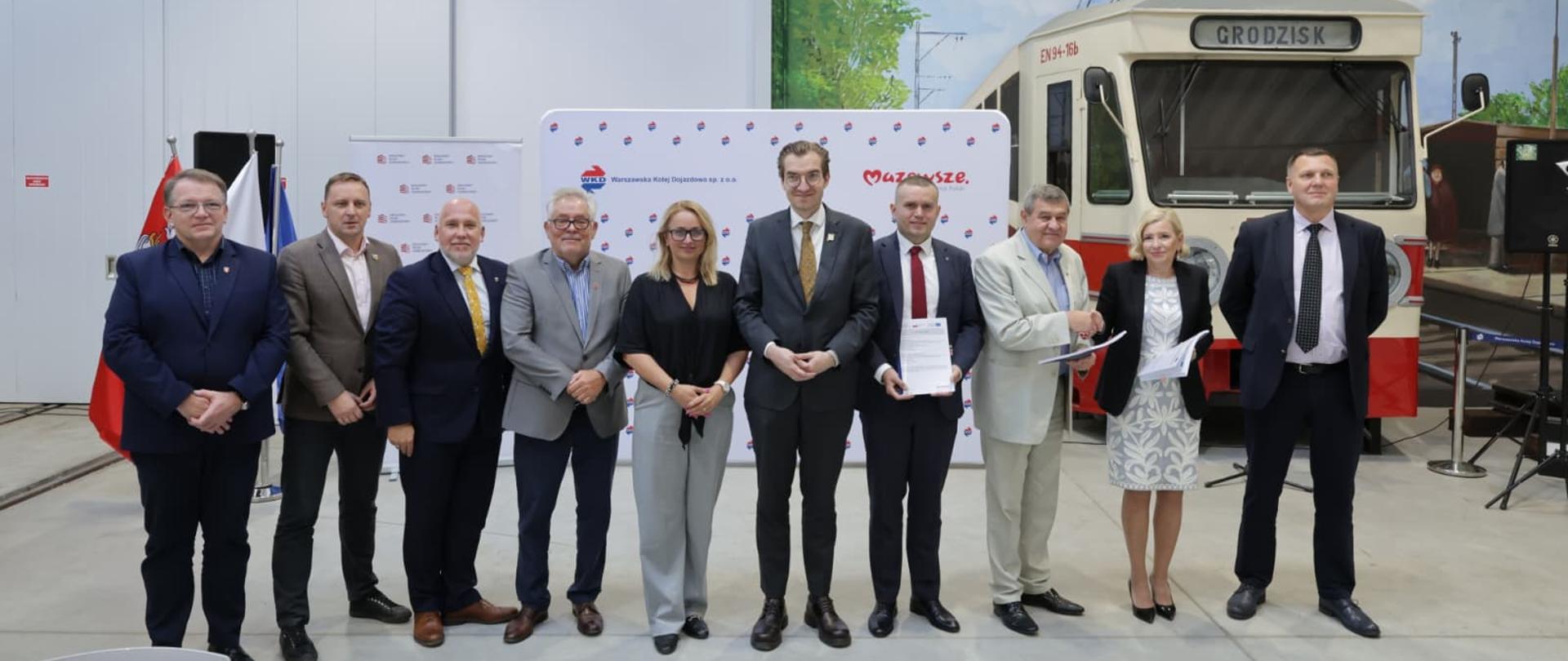 Grupa osób stoi w rzędzie na tle pociągu WKD. Wśród nich wiceminister Jan Szyszko.