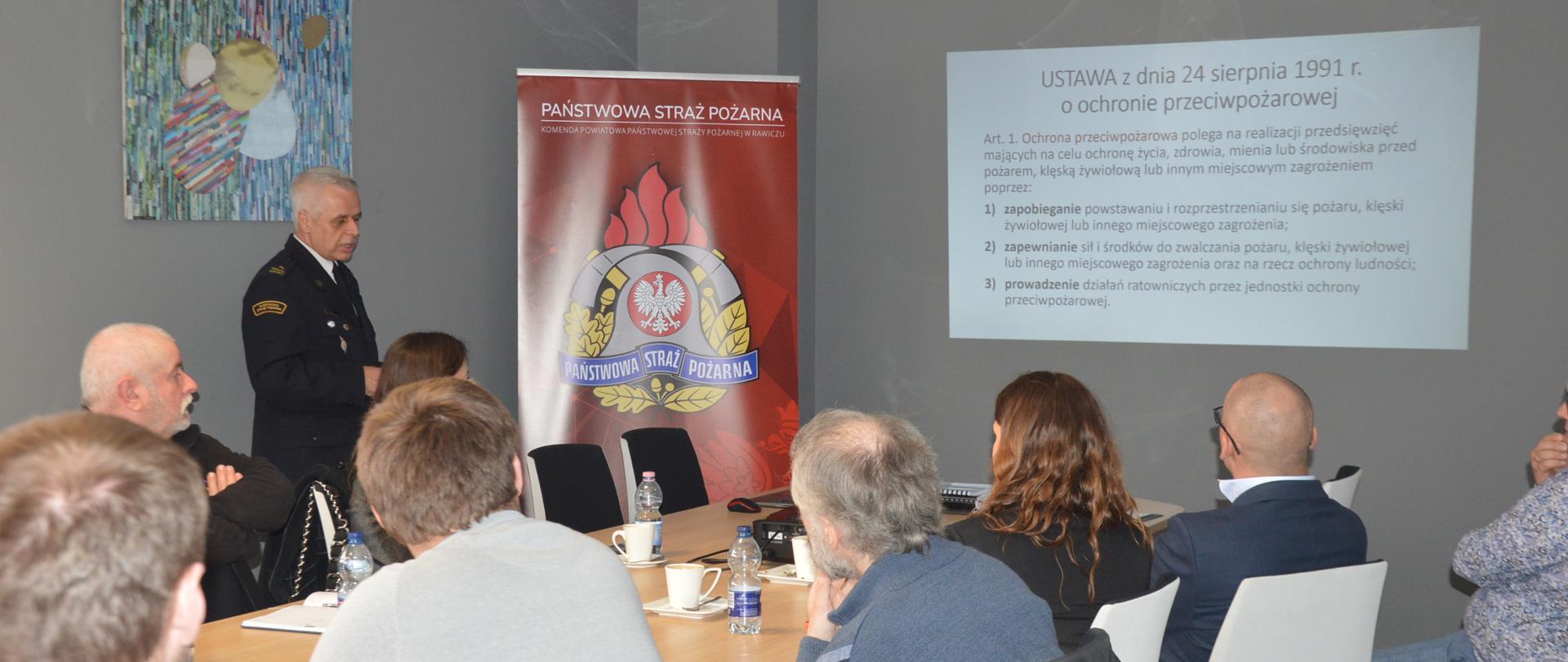 Dzień Bezpieczeństwa i Ochrony Zdrowia w Pracy. Sala konferencyjna Ferm Drobiu Woźniak w Żylicach. W niej uczestnicy konferencji - kierownicy poszczególnych działów firmy, a także przedstawiciele Komendy Powiatowej PSP w Rawiczu, Powiatowej Stacji Sanitarno-Epidemiologicznej w Rawiczu i Państwowej Inspekcji Pracy. Na ścianie wyświetlana jest prezentacja multimedialna. W lewym narożniku pomieszczenia stoi
roll-up promujący Państwową Straż Pożarną.