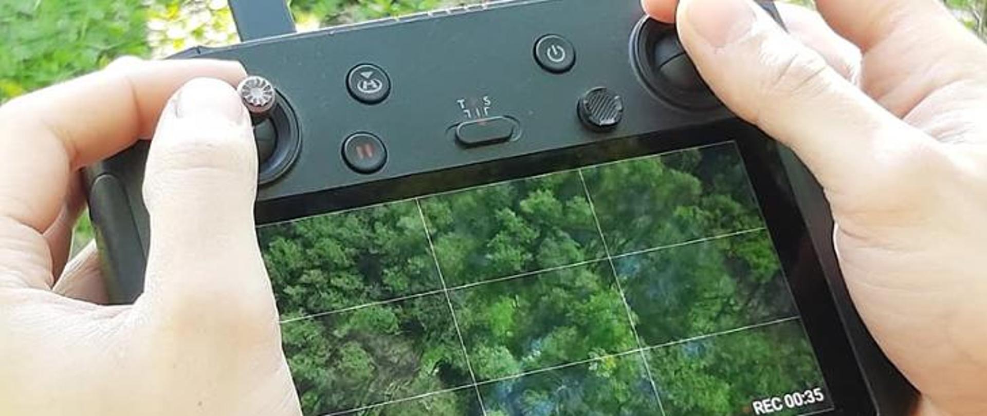 W dużym przybliżeniu widoczne dłonie trzymające kamerę. W niej widać tereny zielone. 