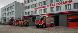 Na zdjęciu widać strażaków w ubraniach bojowych stojących obok samochodów strażackich, samochody stoją przed budynkiem komendy 