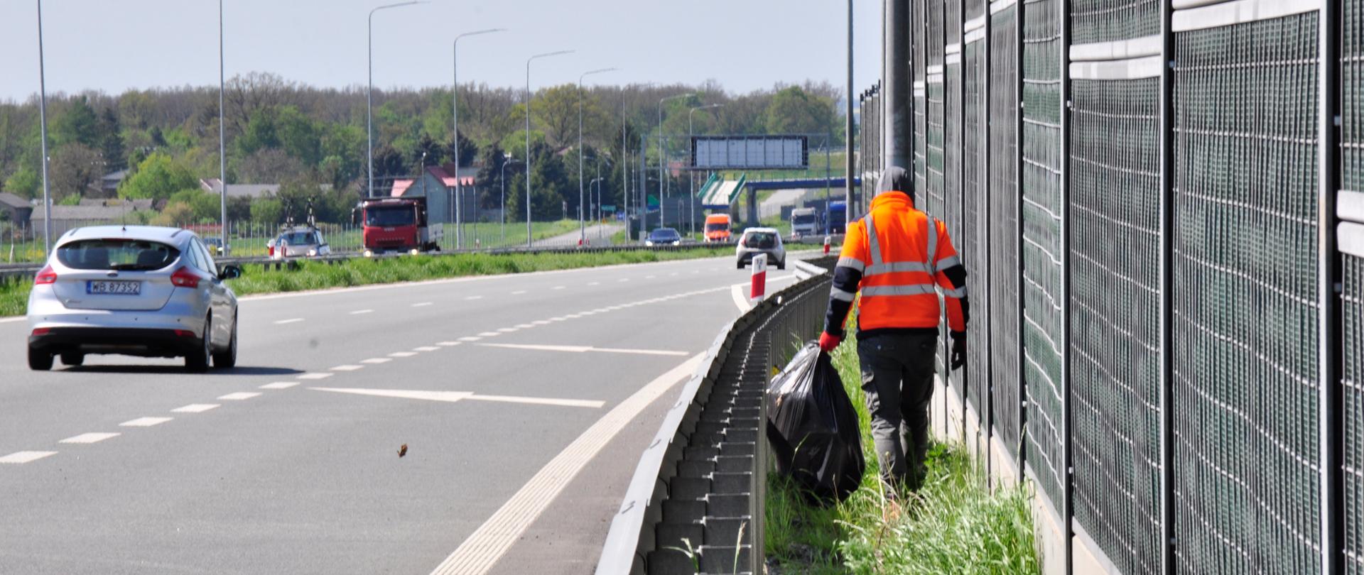 Zdjęcie przedstawia osobę ubraną w pomarańczową kamizelkę odblaskową, która porusza się wzdłuż pobocza autostrady, zbierając śmieci. Osoba ta trzyma duży czarny worek na śmieci i idzie obok metalowego ogrodzenia dźwiękochłonnego. Na drodze w tle widoczne są pojazdy, w tym srebrny samochód osobowy oraz czerwona ciężarówka. Po lewej stronie zdjęcia znajduje się pas zieleni oddzielający jezdnię od pobocza. W tle widoczne są drzewa oraz budynki, a dzień jest pogodny, z lekkim zachmurzeniem.