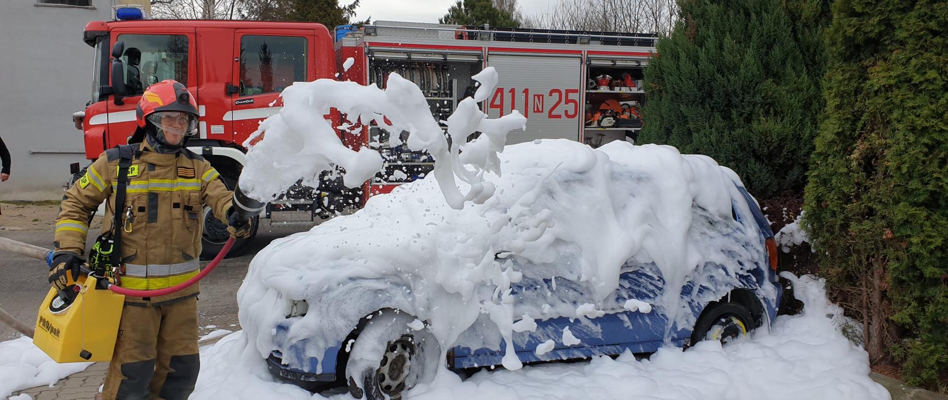 Strażak w hełmie podaje piane na niebieskie auto, ćwiczenia. Za nim w tle duży samochód strażacki.