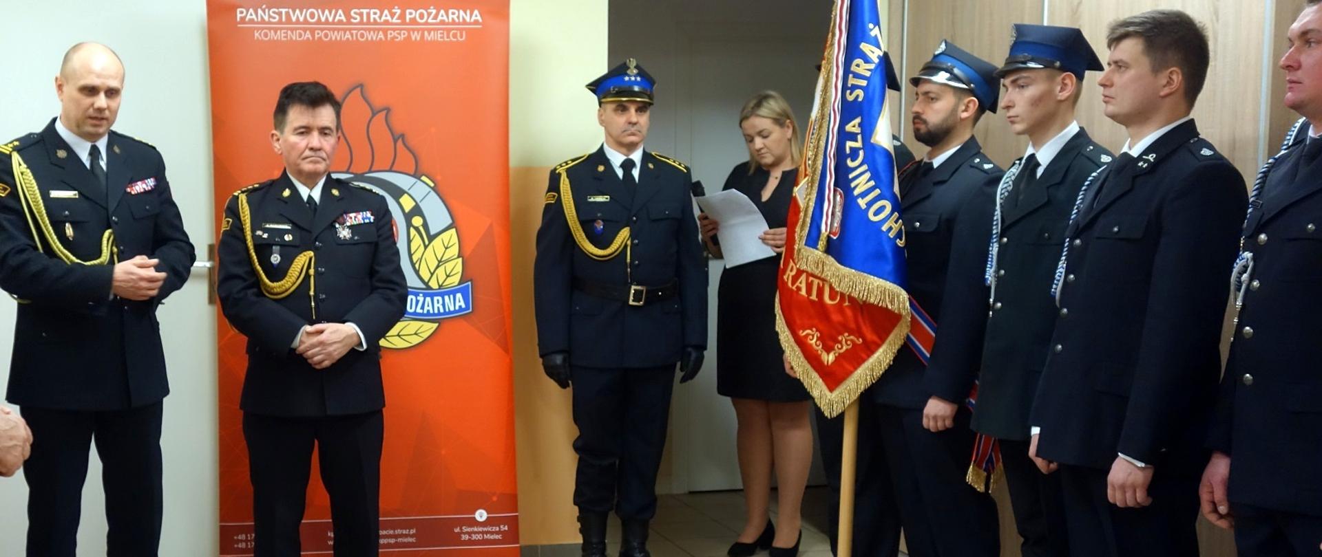 Fotografia wykonana podczas uroczystości włączenia jednostki Ochotniczej Straży Pożarnej Grochowe I do Krajowego Systemu Ratowniczo-Gaśniczego. Na zdjęciu wykonanym w świetlicy jednostki stoją funkcjonariusze Państwowej Straży Pożarnej wraz z druhami Ochotniczej Straży Pożarnej ubrani w mundury galowe koloru granatowego. Są też inni zaproszeni goście i sztandar jednostki włączonej w tym dniu do Krajowego Systemu Ratowniczo Gaśniczego. W tle widać baner z logo i napisem Państwowa Straż Pożarna.