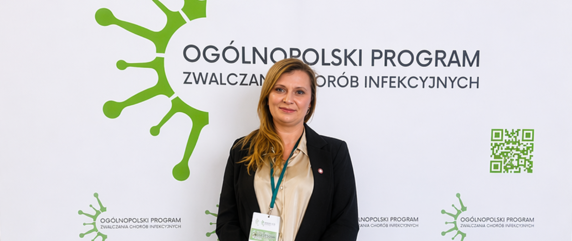 Marta Wiśniewska stoi na tle ścianki promocyjnej z napisem „Ogólnopolski Program Zwalczania Chorób Infekcyjnych”. Po prawej stronie widoczny jest zielony kod QR, a w tle zielone elementy graficzne przypominające strukturę wirusa.
