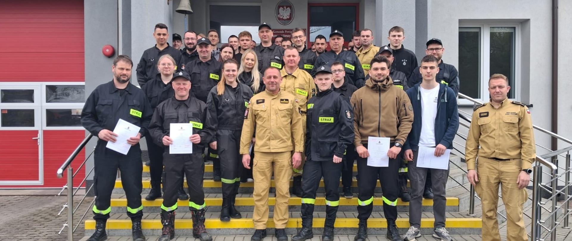 Na zdjęciu widoczna jest liczna grupa strażaków OSP, która stoi na schodach przed budynkiem. Na zdjęciu widocznych jest dwóch funkcjonariuszy w piaskowych mundurach. Część strażaków z OSP trzyma dokumenty.