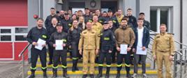 Na zdjęciu widoczna jest liczna grupa strażaków OSP, która stoi na schodach przed budynkiem. Na zdjęciu widocznych jest dwóch funkcjonariuszy w piaskowych mundurach. Część strażaków z OSP trzyma dokumenty.