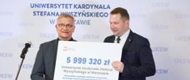 Minister Czarnek stoi obok starszego mężczyzny w garniturze, trzymają duży symboliczny czek z napisem 5 999 320 zł, za nimi na ścianie napis UKSW.