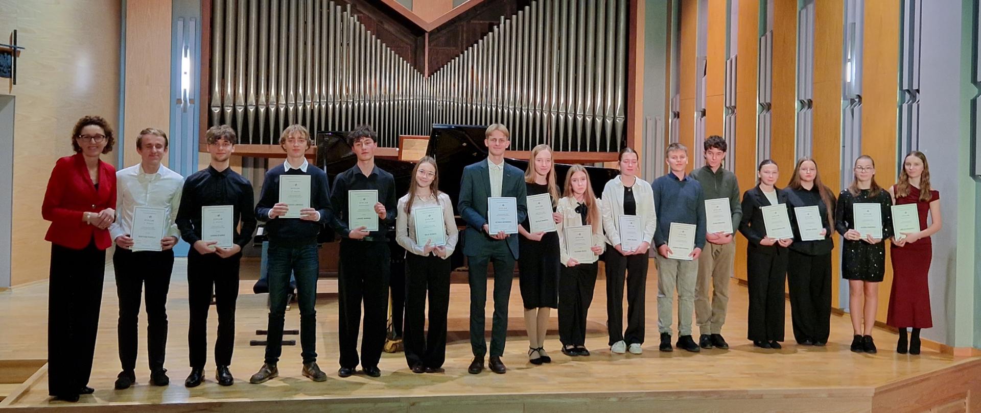 Zdjęcie przedstawia grupę szesnastu osób stojących w rzędzie na scenie sali koncertowej. Jest to uroczystość wręczenia dyplomów lub zakończenie konkursu muzycznego. Uczestnicy: W rzędzie stoi piętnaście młodych osób (dziewcząt i chłopców) oraz jedna dorosła kobieta po lewej stronie. Ubiór: Dominują stroje wizytowe w ciemnych kolorach (czernie, granaty), biele oraz okazjonalnie czerwień i zieleń. Większość młodych ludzi trzyma przed sobą białe arkusze papieru – dyplomy lub certyfikaty. Ustawienie: Grupa stoi przodem do widza, uśmiechając się lub zachowując powagę. Instrumenty: Bezpośrednio za grupą, w centralnym punkcie, znajduje się czarny fortepian skrzydłowy. Nad nim górują monumentalne, srebrne piszczałki organów ułożone w kształt litery "M" lub rozłożystych skrzydeł. Wnętrze: Sala ma nowoczesny wystrój. Ściany są wyłożone jasnymi, drewnianymi panelami przeplatanymi szarymi elementami. Na suficie widać punktowe oświetlenie. Perspektywa: Zdjęcie wykonano z poziomu widowni; w dolnej części kadru widoczne są oparcia pustych, granatowych foteli kinowych/teatralnych.