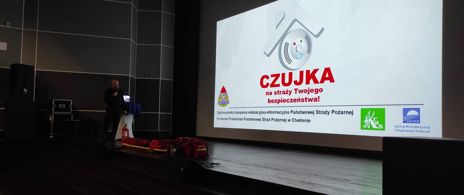 Na zdjęciu ml. kpt. Kasper Korczak podczas prowadzenia wykładu w Kinoteatrze Rondo w Chełmnie, dodatkowo przed ekranem kinowym znajdują się torba medyczna PSP R1, oraz nosze typu deska.