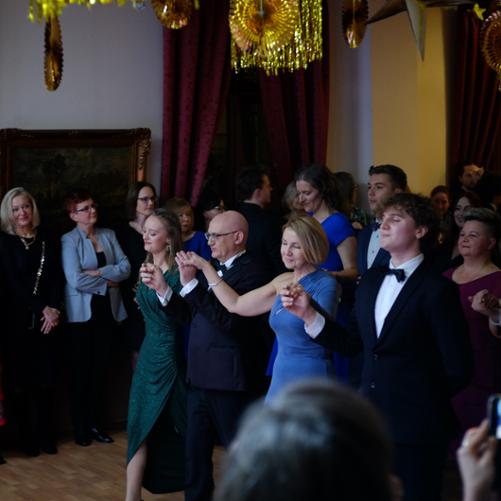 zdjęcie przedstawia dyrekcję szkoły, wychowawczynię klasy oraz uczniów i uczennice klasy maturalnej podczas innej figury Poloneza na sali bankietowe,j w tle inni nauczyciele obserwujący taniec