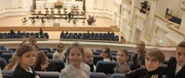 Dzieci w fotelach widowni, koloru granatowego, obrócone tyłem do sceny, tło stanowi scena na której stoją krzesła i pulpity na nuty. Po prawej stronie sceny stoją dwa instrumenty