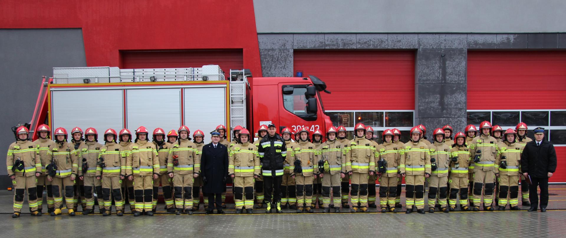 Pamiątkowe zdjęcie: w kadrze umieszczono ratowników z Jednostki Ratowniczo-Gaśniczej nr 1 w Kielcach wraz z Komendantem Miejskim PSP, zastępcą dowódcy Jednostki Ratowniczo-Gaśniczej nr 1 i zastępcą naczelnika wydziału organizacyjno-kadrowego KM PSP na tle samochodu ratownictwa technicznego. Na dalszym planie nowa siedziba JRG nr 1 w Kielcach.
