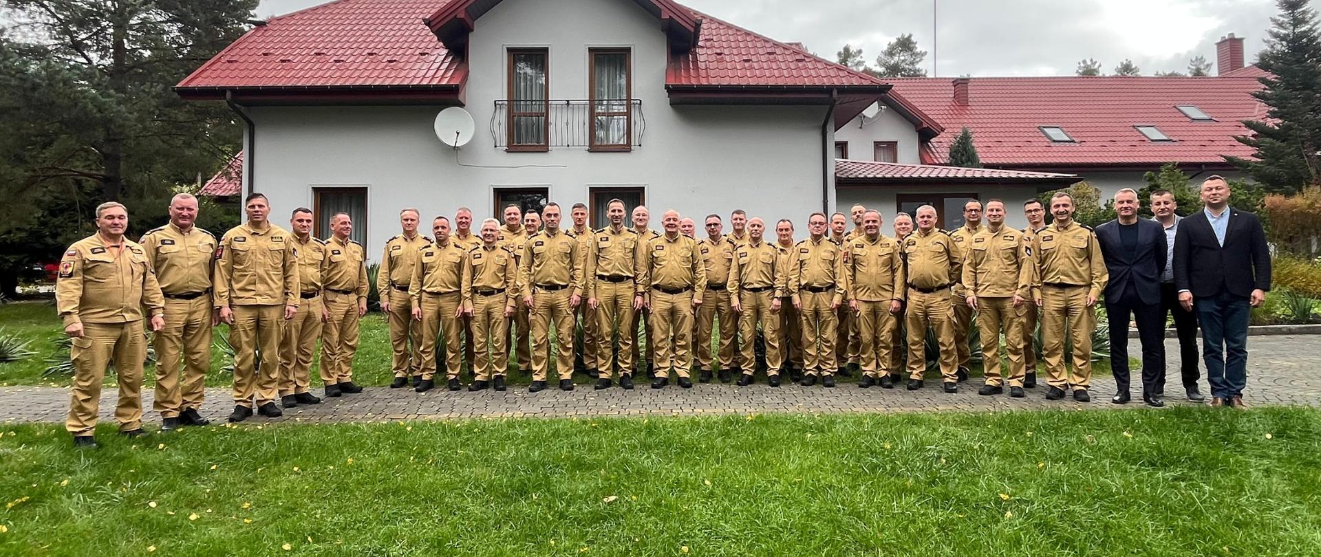 Duża grupa mężczyzn, w większości ubranych w mundury służbowe, stoi w rzędzie na trawniku przed dużym budynkiem z czerwonym dachem, pozując do wspólnego zdjęcia. Kilku mężczyzn po prawej stronie jest ubranych w garnitury.