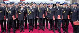 Grupowe zdjęcie funkcjonariuszy PSP z województwa lubuskiego w galowych mundurach, wykonane po uroczystej promocji na dziedzińcu Szkoły Aspirantów PSP w Poznaniu. Strażacy stoją na czerwonym dywanie, trzymając w dłoniach czerwone teczki z godłem Polski – dokumenty potwierdzające uzyskanie tytułu technika pożarnictwa. W tle widoczne biało-czerwone namioty oraz transparent Szkoły Aspirantów.