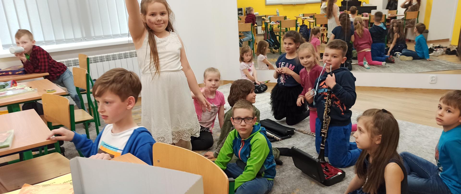 Na dywanie w klasie siedzą, klęćzą i stoją kolorowo ubrane dzieci, lustro na ścianie odbija ich postaci od tyłu. Dzieci patrzą się w różnych kierunkach, a jedno z nich wyjmuje klarnet z futerału. Ściana jest żółta, ławki z jasnego drewna z zielonymi nóżkami.
