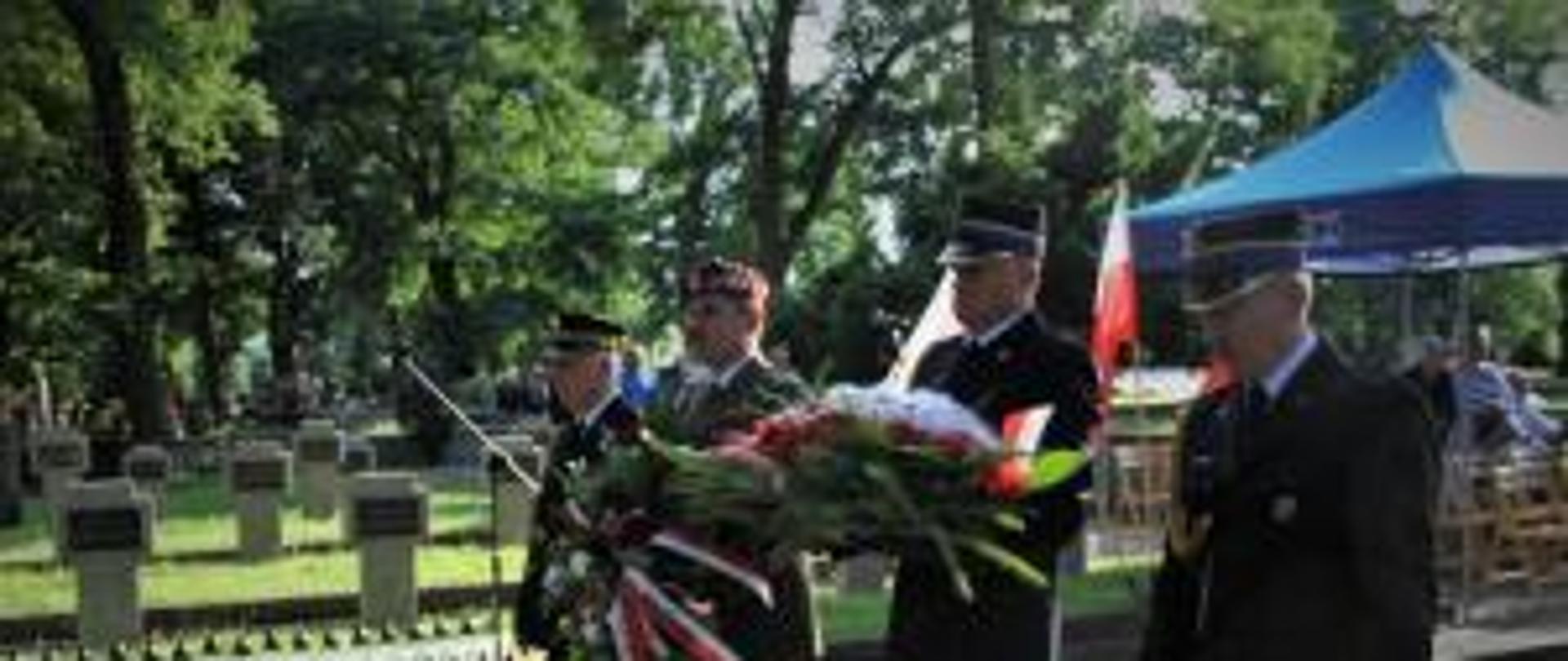 Czterej mężczyźni w mundurach idą z kwiatami w rękach. W tle widać drzewa i niebieski namiot. 