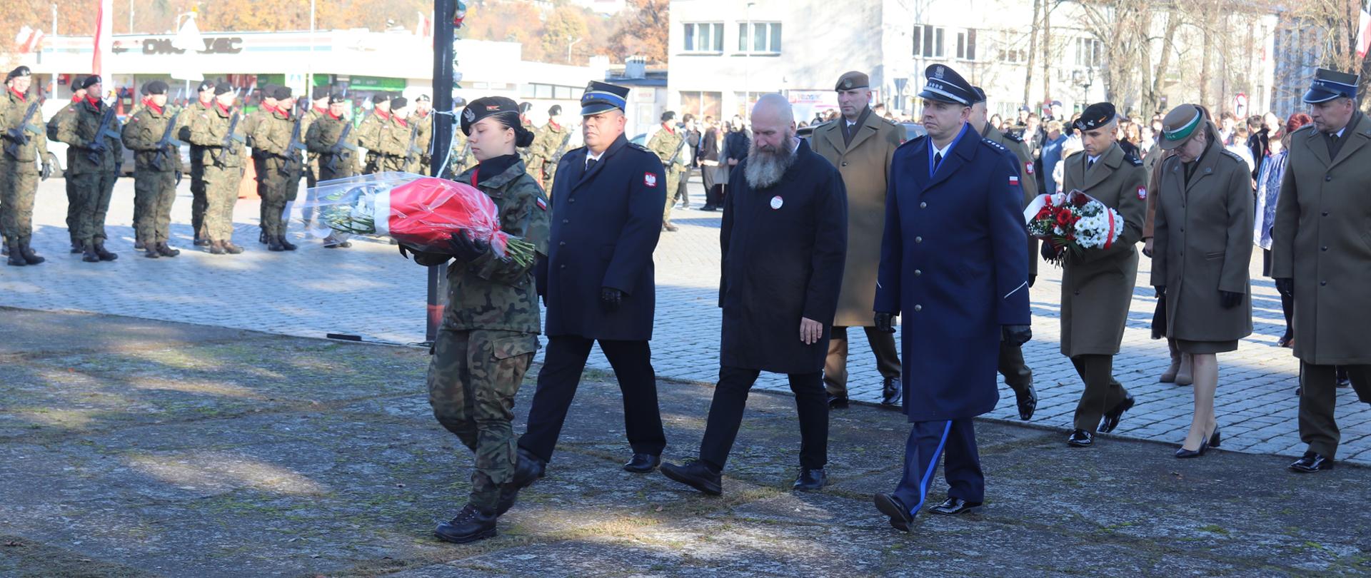 zdjęcie przedstawia żołnierza Wojska Polskiego niosącego kwiaty pod pomnik, następnie znajdują się przedstawiciele służb mundurowych.