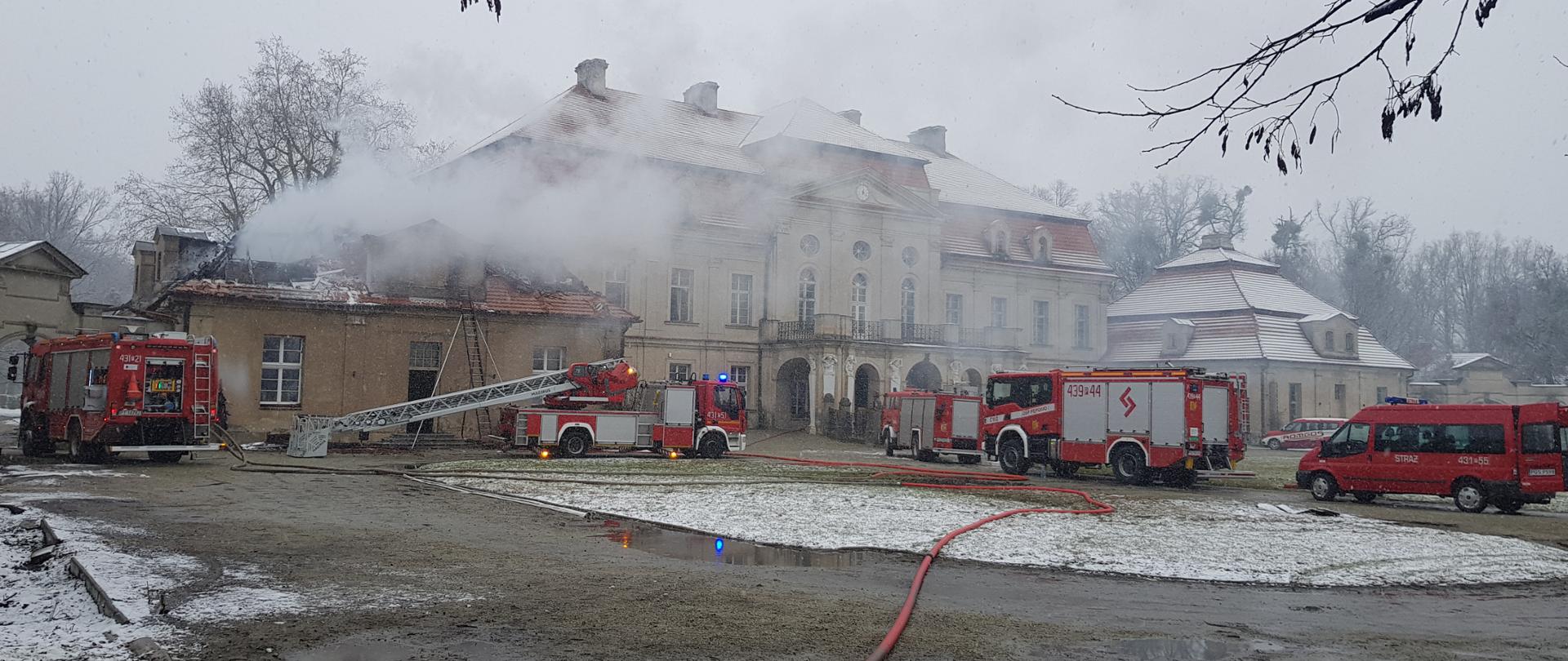 Zdjęcie przedstawia pożaru pałacu w fazie dogaszania. Na zdjęcie znajduje się 5 samochodów strażackich. Strażacy prowadza prace rozbiórkowe. Dach alkierza uległ spaleniu.