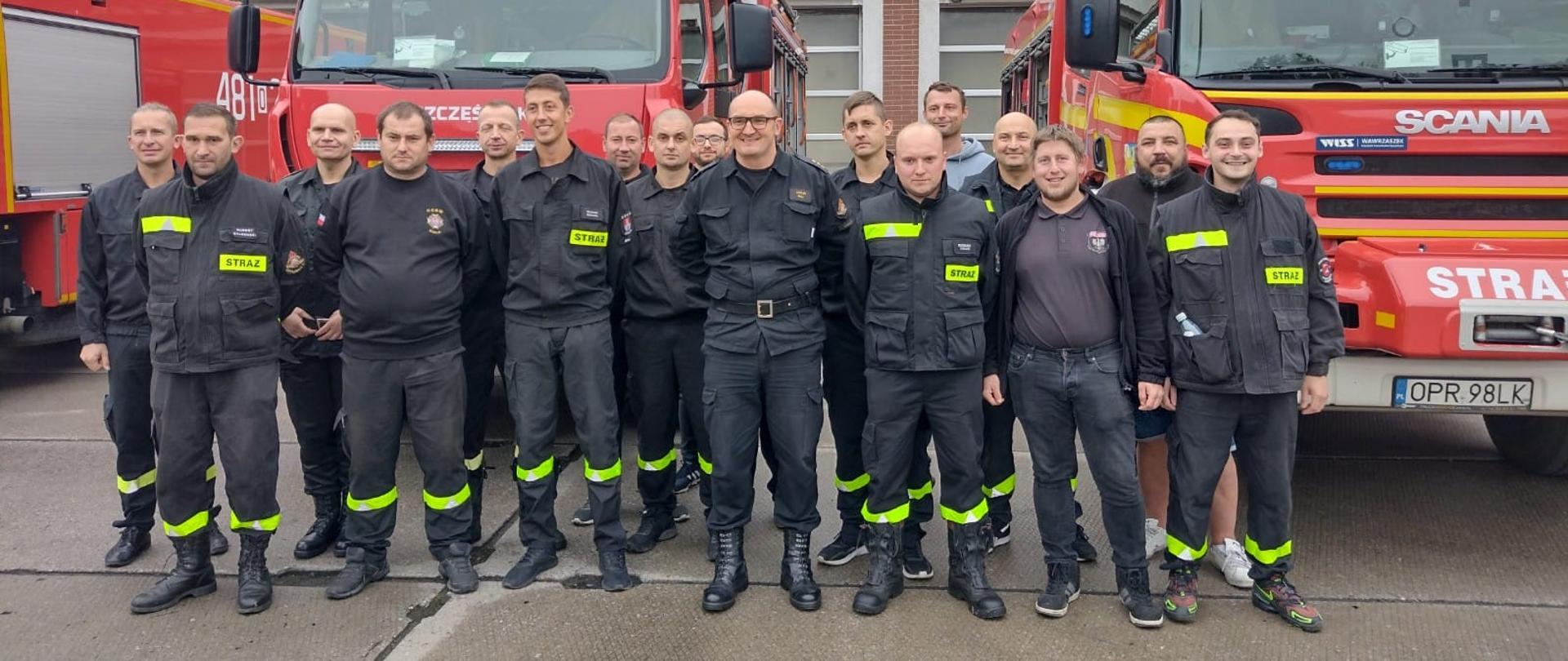 Zdjęcie przedstawia strażaków w mundurach i ubraniach cywilnych na placu Komendy Powiatowej PSP. W tle samochody pożarnicze i budynek strażnicy.
samochód pożarniczy i budynek strażnicy.