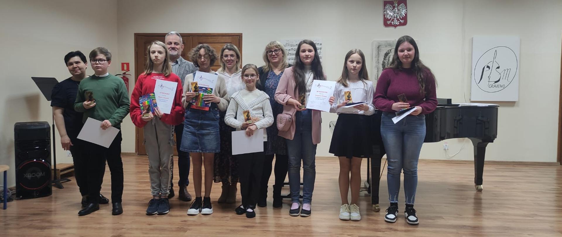 Zdjęcie przedstawia grupę uczniów, którzy zostali wyróżnieni w VII Szkolnym Konkursie ,,Mistrz Dyktanda". Dzieci stoją z dyplomami i statuetkami. Za nimi stoi pan Bogusław Rząsa oraz nauczyciele, którzy zorganizowali konkurs (Joanna Jabłońska, Ewelina Chrzanowska i Urszula Właźniak). W tle fortepian, godło, głośnik multimedialny oraz grafiki w barwach biało czarnych. 