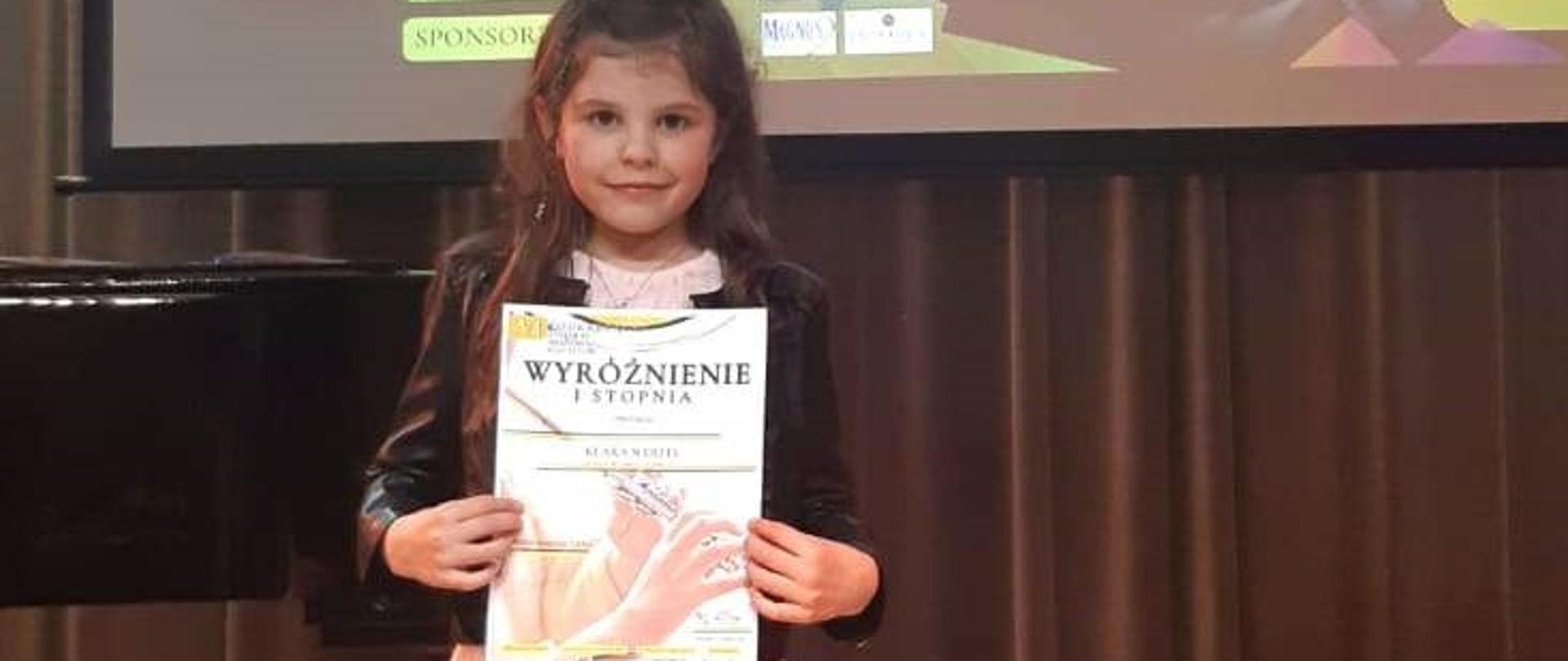 Uczennica trzymająca dyplom wyróżnienia I stopnia. Z tyłu widać fortepian.