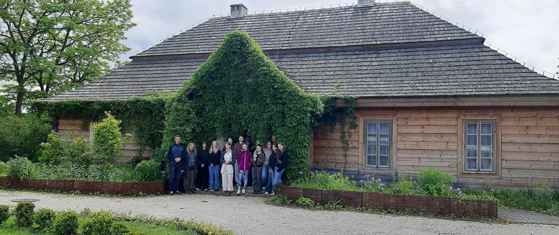 Na tle drewnianego budynku pozuje kilkanaście osób. Wejście do budynku i ściana są porośnięte bluszczem. Przed budynkiem rosną krzewy i kwiaty, stoi wiklinowy płotek, jest wyżwirowana ścieżka.
