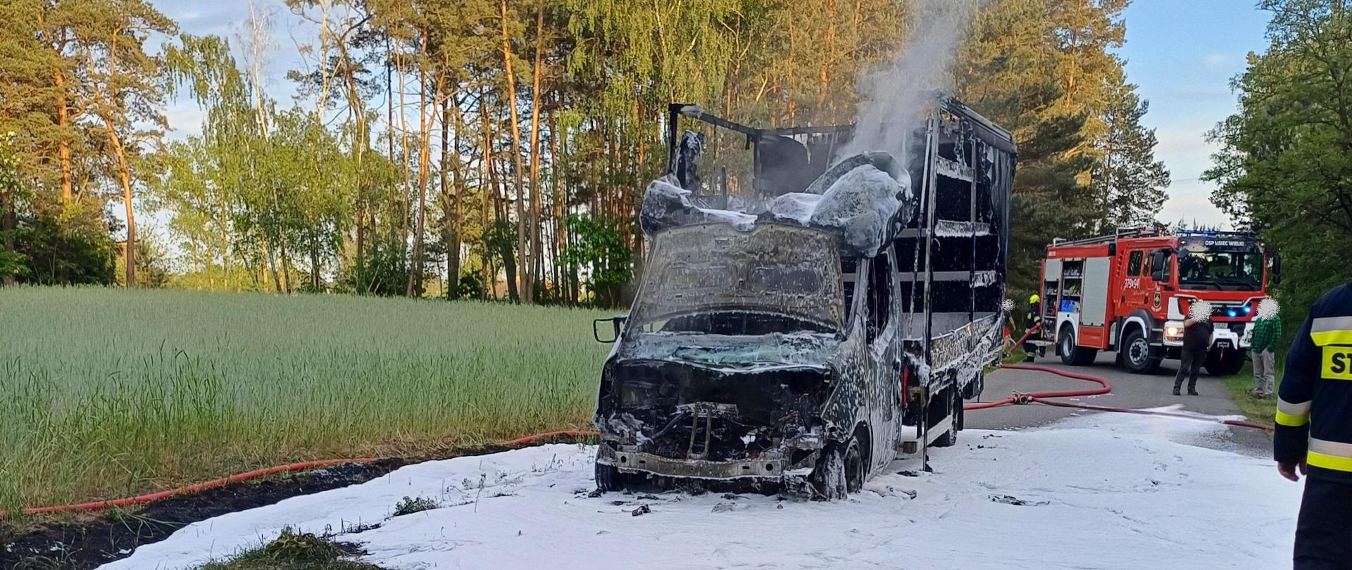 Zdjęcia przedstawia spalony samochód dostawczy marki Renault. Nad samochodem unosi się dym. Z tyłu widać samochód strażacki. Pod samochodem oraz na jezdni widać pianę gaśniczą oraz dwie linie wężowe. 