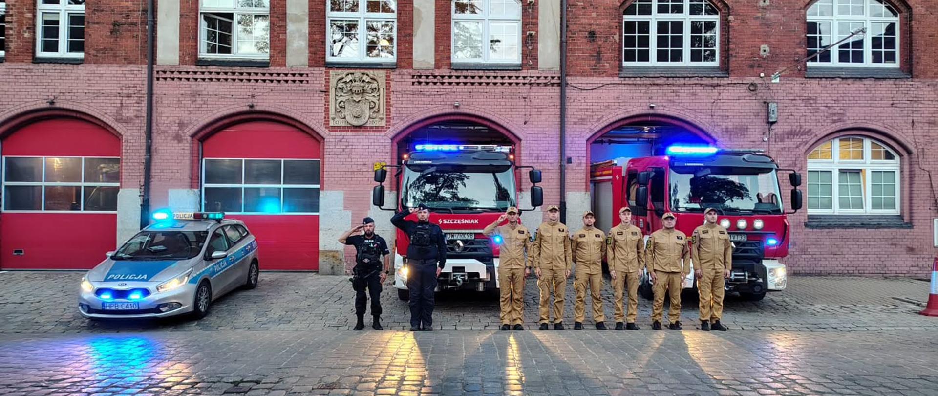 Przed budynkiem z czerwonej cegły stoją w szeregu dwaj funkcjonariusze Policji w czarnych mundurach oraz siedmiu strażaków w piaskowych mundurach służbowych. Niektórzy z nich salutują. W tle widoczne są dwa czerwone wozy strażackie z włączonymi niebieskimi sygnałami świetlnymi oraz oznakowany radiowóz policyjny