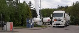 Na zdjęciu biała cysterna do transportu paliwa. Obok biały zbiornik propan-butan z którego wydobywa się biały obłok. Zdjęcie zrobione w ciągu dnia na stacji benzynowej. W okolicach drzewa, zielone. Zachmurzenie duże.