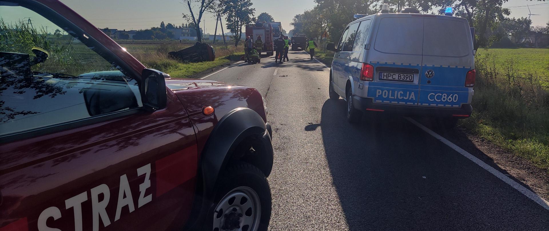 Zdjęcie przedstawia pas drogowy drogi krajowej nr 62, na której znajdują się pojazdy służb ratowniczych (dwa wozy straży pożarnej, policja) strażaków i policjantów podczas czynności służbowych po kolizji drogowej pojazdu osobowego z zestawem rolniczym w postaci ciągnika oraz dwóch przyczep.