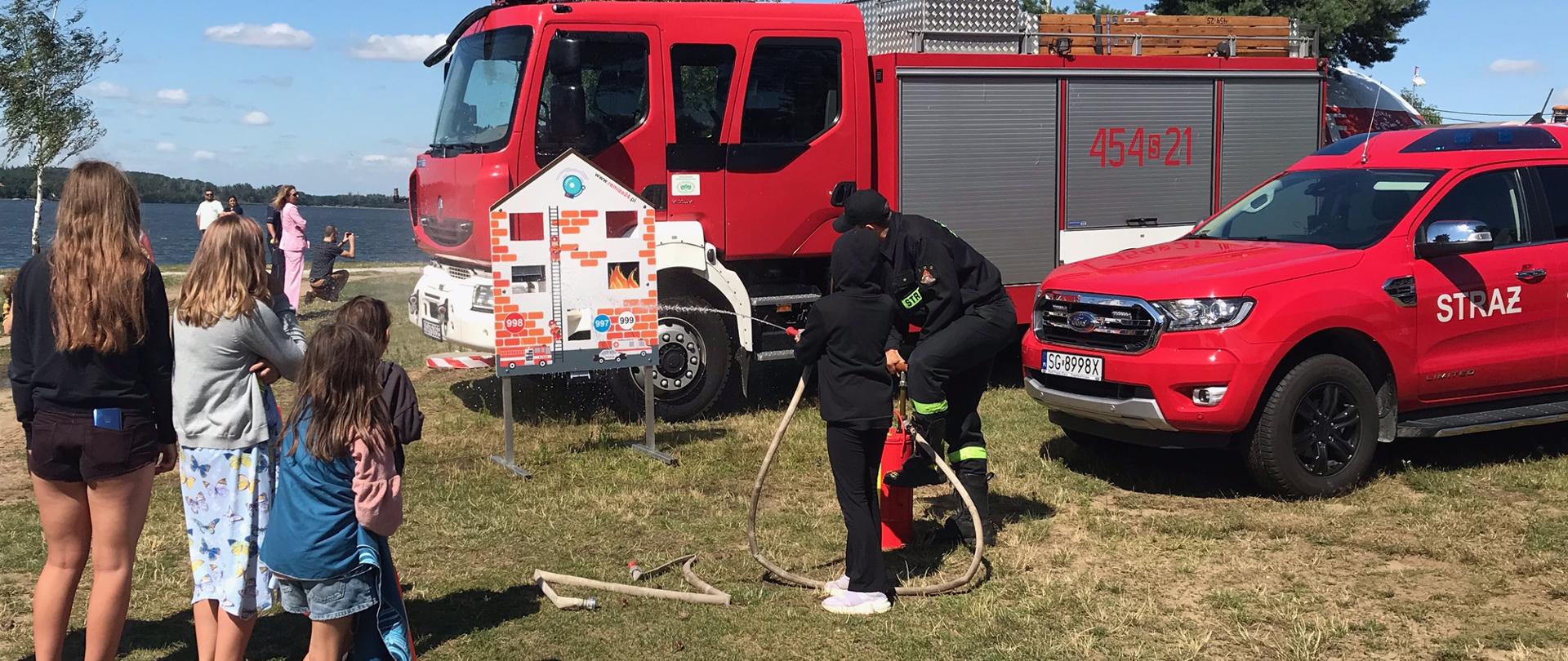 dziecko razem ze strażakiem celuje prądem wody w domek z planszę w kształcie domku z ognikami do strącania, obok przygląda się czwórka dzieci, w te samochód ratowniczo - gaśniczy, obok czerwony samochód terenowy
