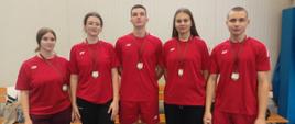 Na zdjęciu stoi pięcioro młodych sportowców w czerwonych koszulkach i z medalami na szyjach. Ustawieni są w jednym rzędzie, na tle jasnej ściany sali gimnastycznej.