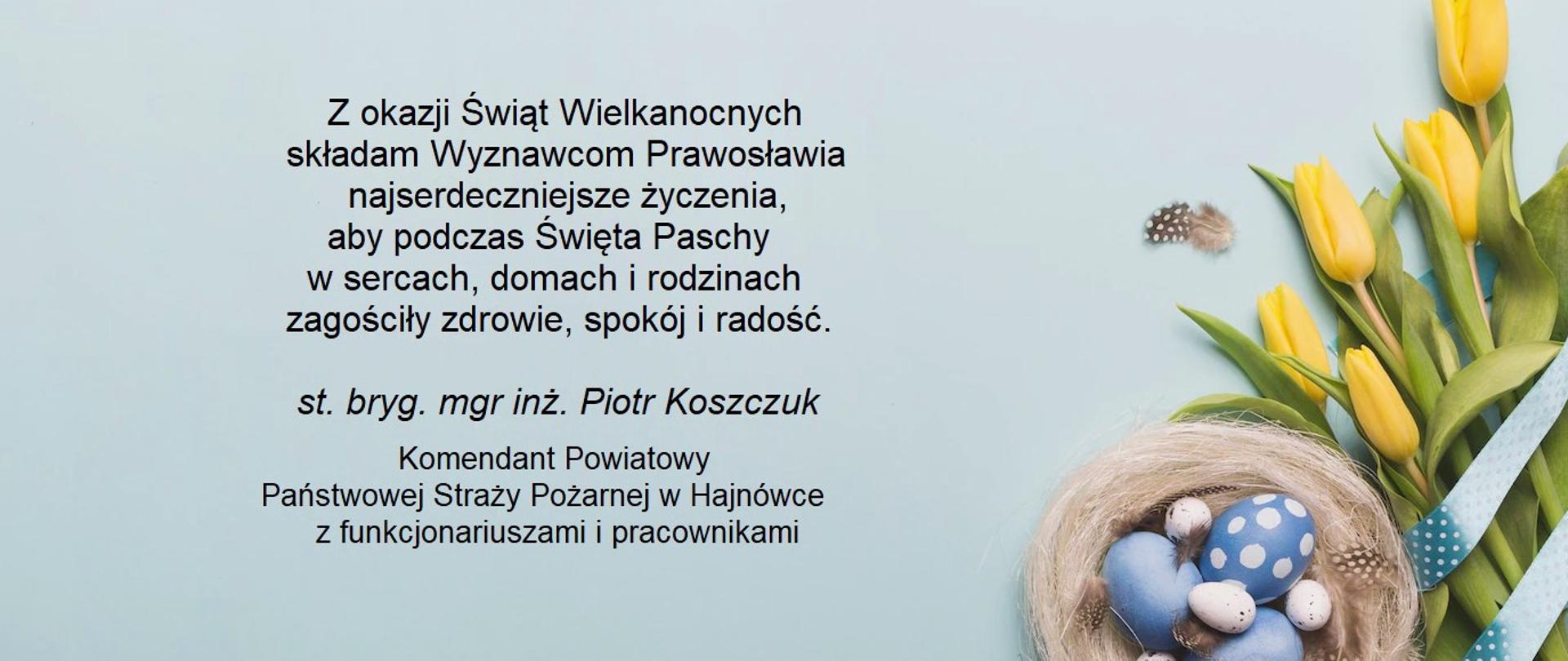 Na jasnoniebieskim tle z prawej strony bukiet kwiatów z malowanymi na niebiesko i szaro jajkami. Jajka duże i malutkie leżą w jasnobrązowym gniazdku. Po lewej stronie życzenia z okazji świąt Wielkanocnych Komendanta Powiatowego PSP w Hajnówce.