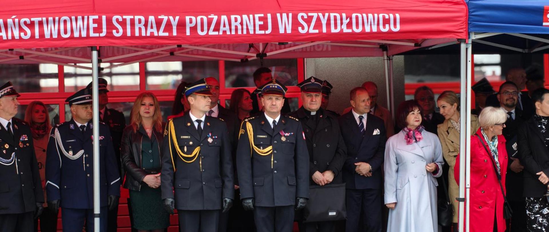 Zdjęcie przedstawia oficjalną uroczystość plenerową z udziałem przedstawicieli służb mundurowych oraz osób cywilnych. Uczestnicy stoją w rzędzie pod zadaszeniem dużego, czerwonego namiotu promocyjnego. Na froncie namiotu widnieje biały, drukowany napis (widoczny we fragmencie): „AŃSTWOWEJ STRAŻY POŻARNEJ W SZYDŁOWCU” (będący częścią pełnej nazwy: Komenda Powiatowa Państwowej Straży Pożarnej w Szydłowcu).