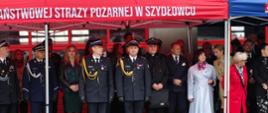 Zdjęcie przedstawia oficjalną uroczystość plenerową z udziałem przedstawicieli służb mundurowych oraz osób cywilnych. Uczestnicy stoją w rzędzie pod zadaszeniem dużego, czerwonego namiotu promocyjnego. Na froncie namiotu widnieje biały, drukowany napis (widoczny we fragmencie): „AŃSTWOWEJ STRAŻY POŻARNEJ W SZYDŁOWCU” (będący częścią pełnej nazwy: Komenda Powiatowa Państwowej Straży Pożarnej w Szydłowcu).