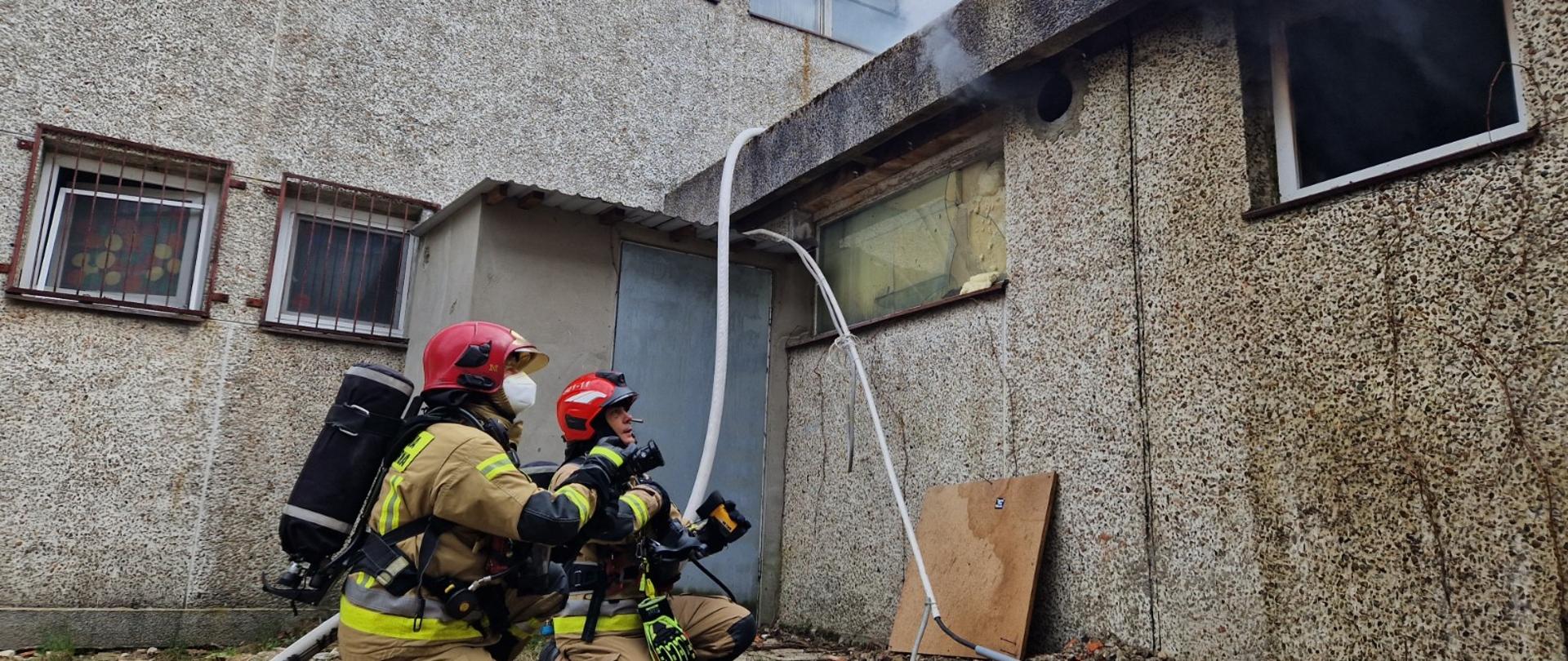 dwóch strażaków klęczy przed budynkiem, wokoło węże z wodą