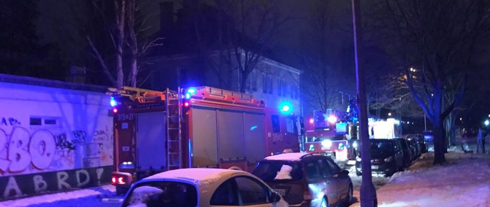 Na pierwszym planie widać zaparkowane samochody, w tym czerwony samochód strażacki i drabinę mechaniczną. Światła samochodów pożarniczych świecą na niebiesko. W tle widoczny jest trzykondygnacyjny budynek i drzewa.