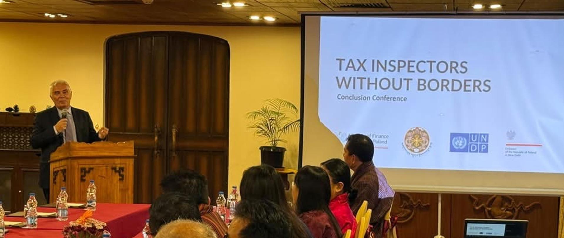 Dr. Piotr Antoni Świtalski delivers remarks during the concluding conference of the Tax Inspectors Without Borders project in Bhutan. Representatives of Bhutanese institutions and project partners are seated around conference tables, while a presentation screen displays the title of the programme.